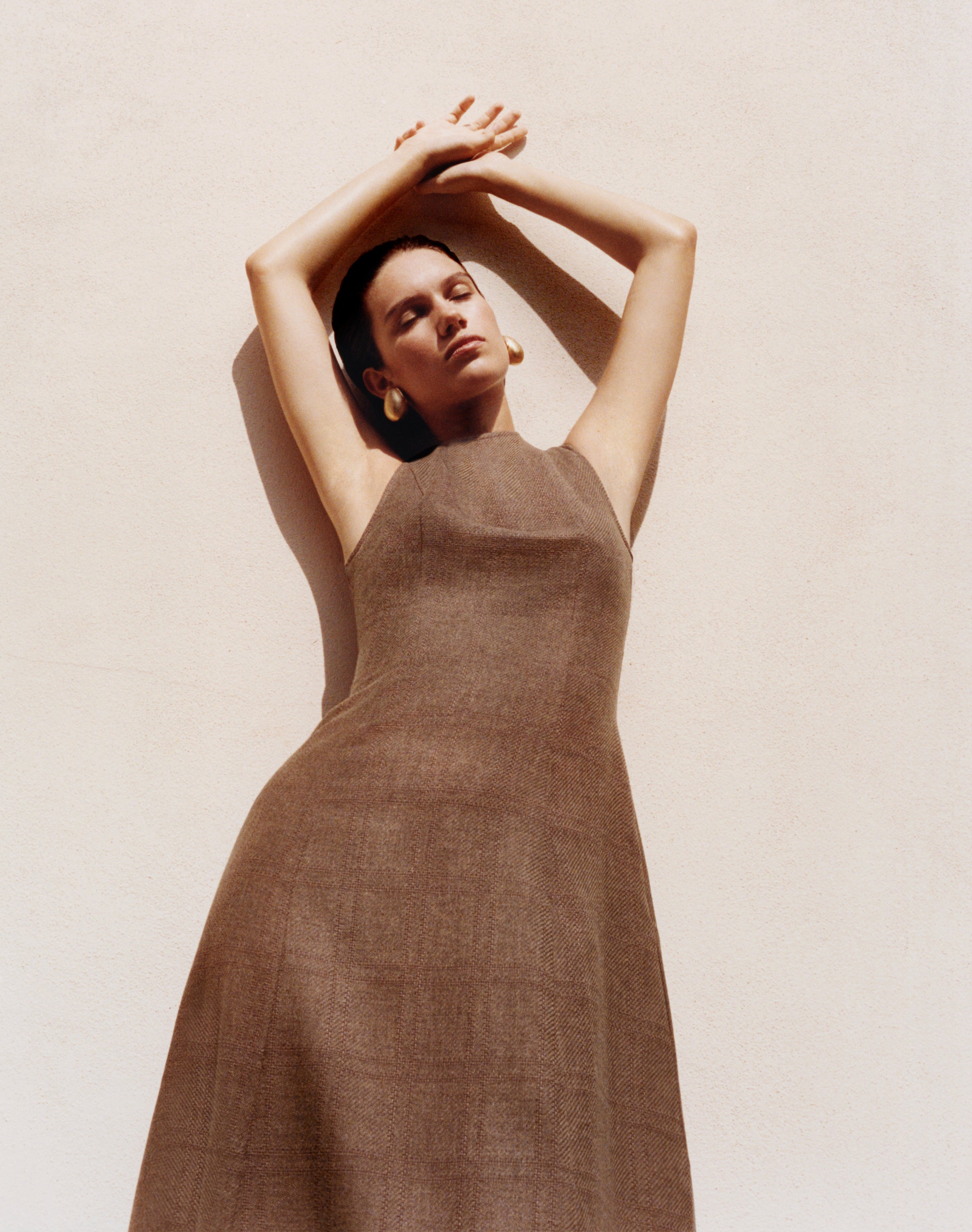 A woman wearing a sleeveless brown linen dress poses with her arms raised against a textured beige wall.