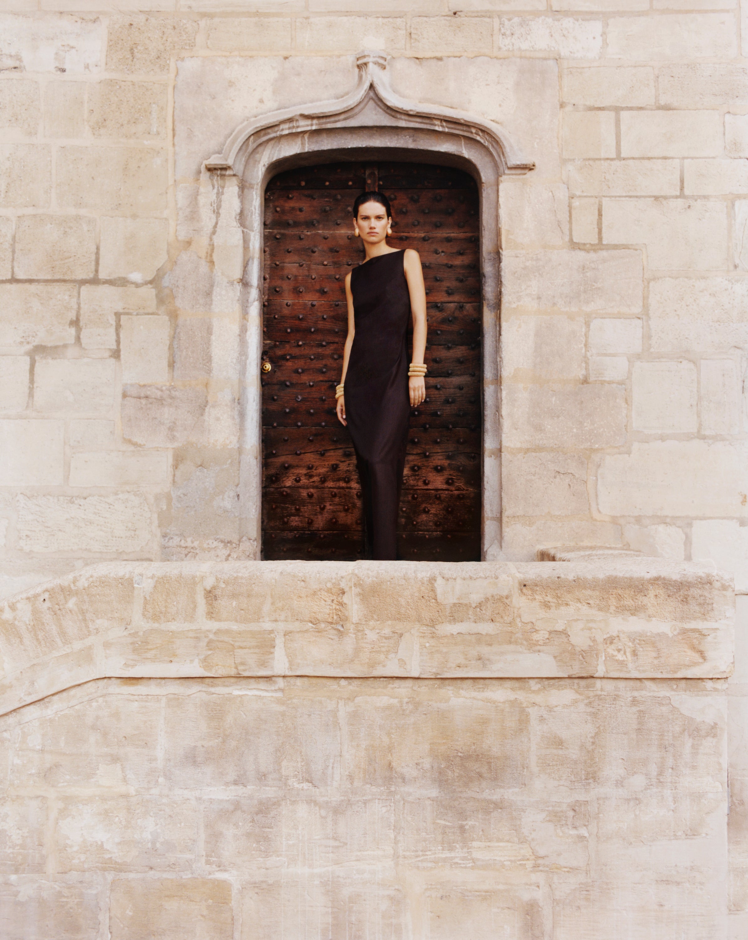 A woman wearing the Yves dress, sleeveless black dress stands in a stone archway doorway with a dark wooden door behind her, set against a beige stone wall.