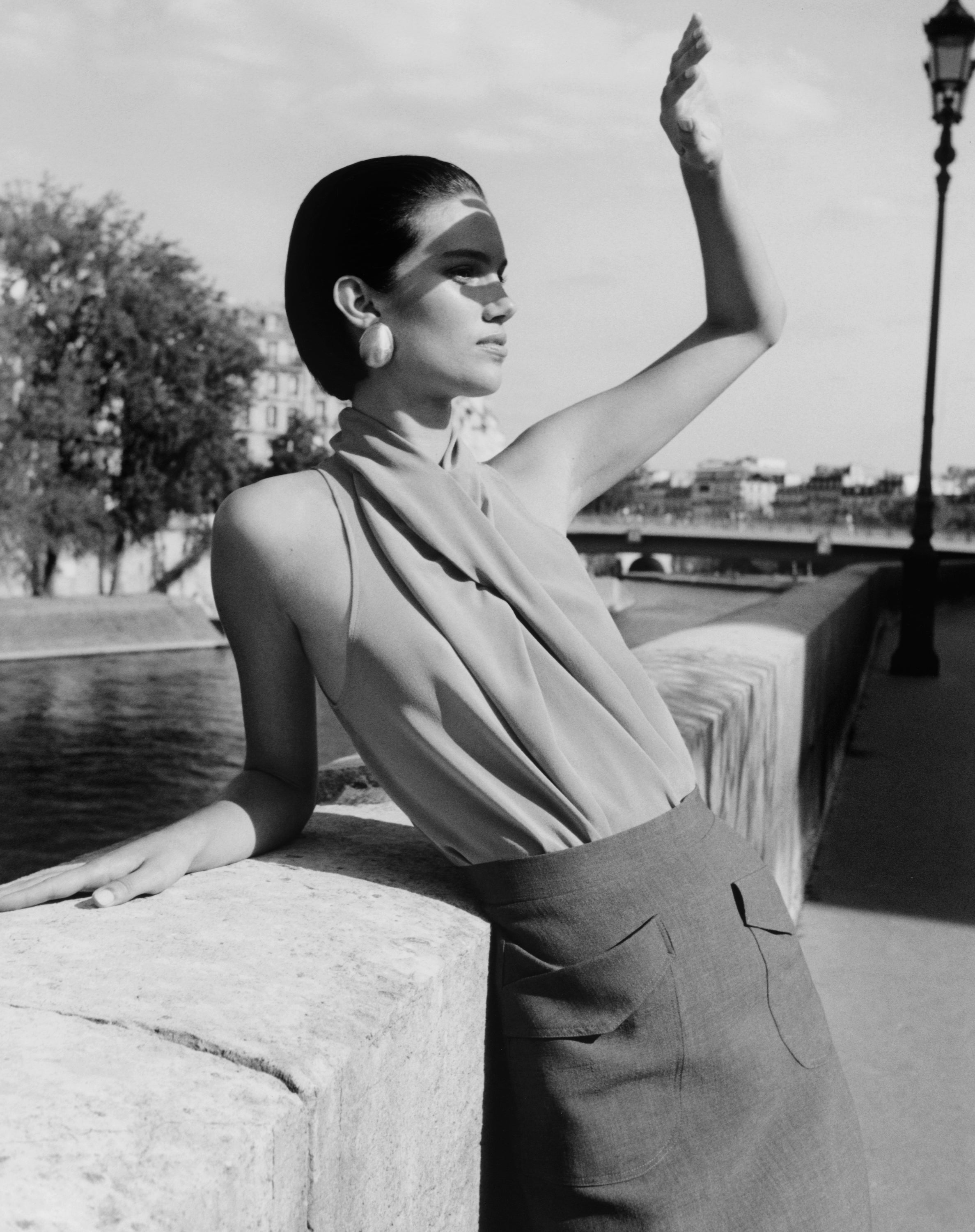 A black and white fashion photograph of a woman leaning on a stone bridge railing next to water, wearing a draped sleeveless top and high-waisted skirt.