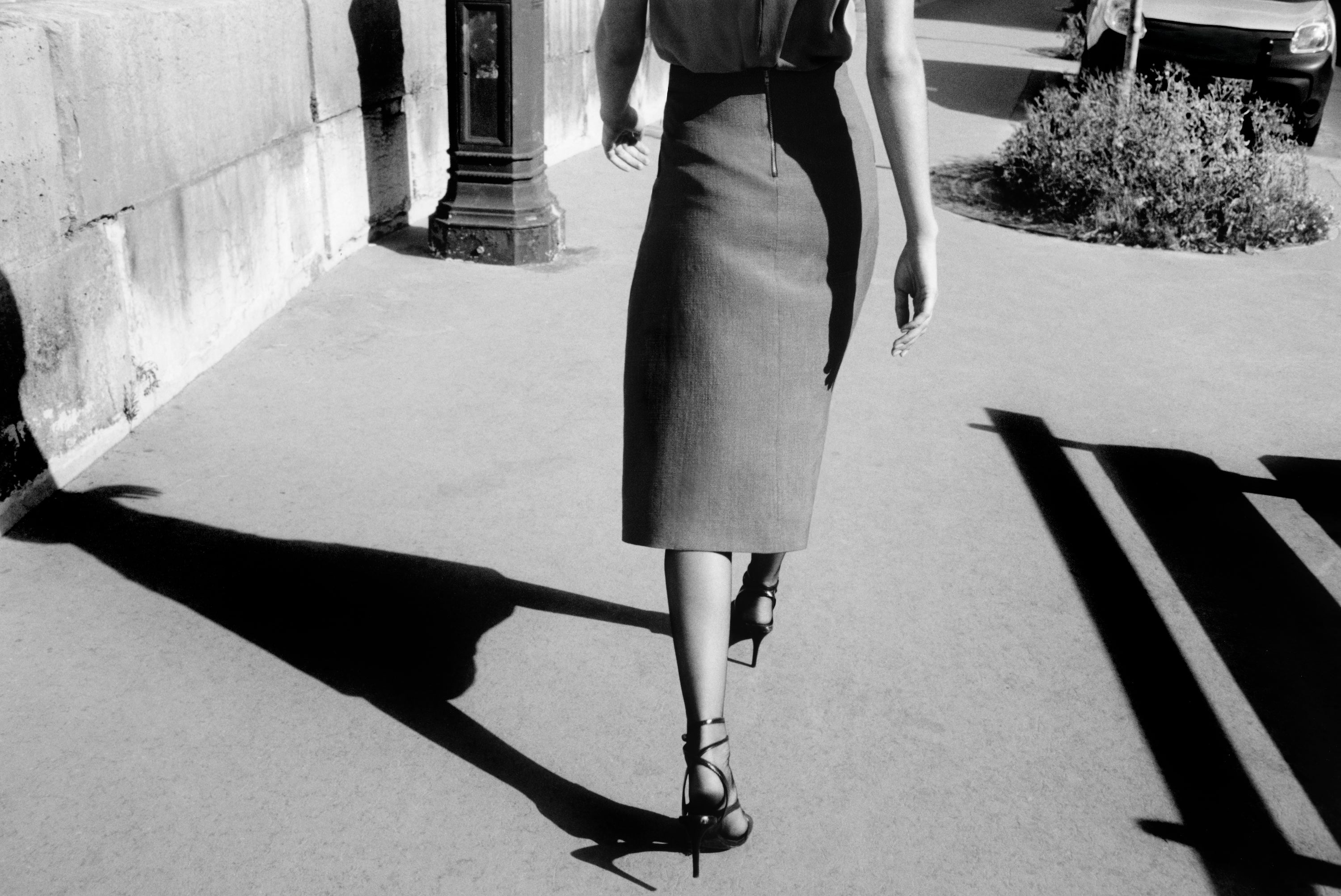 A black and white fashion photograph of a woman from behind walking away on a sunlit sidewalk, wearing a knee-length textured skirt and strappy high heels.