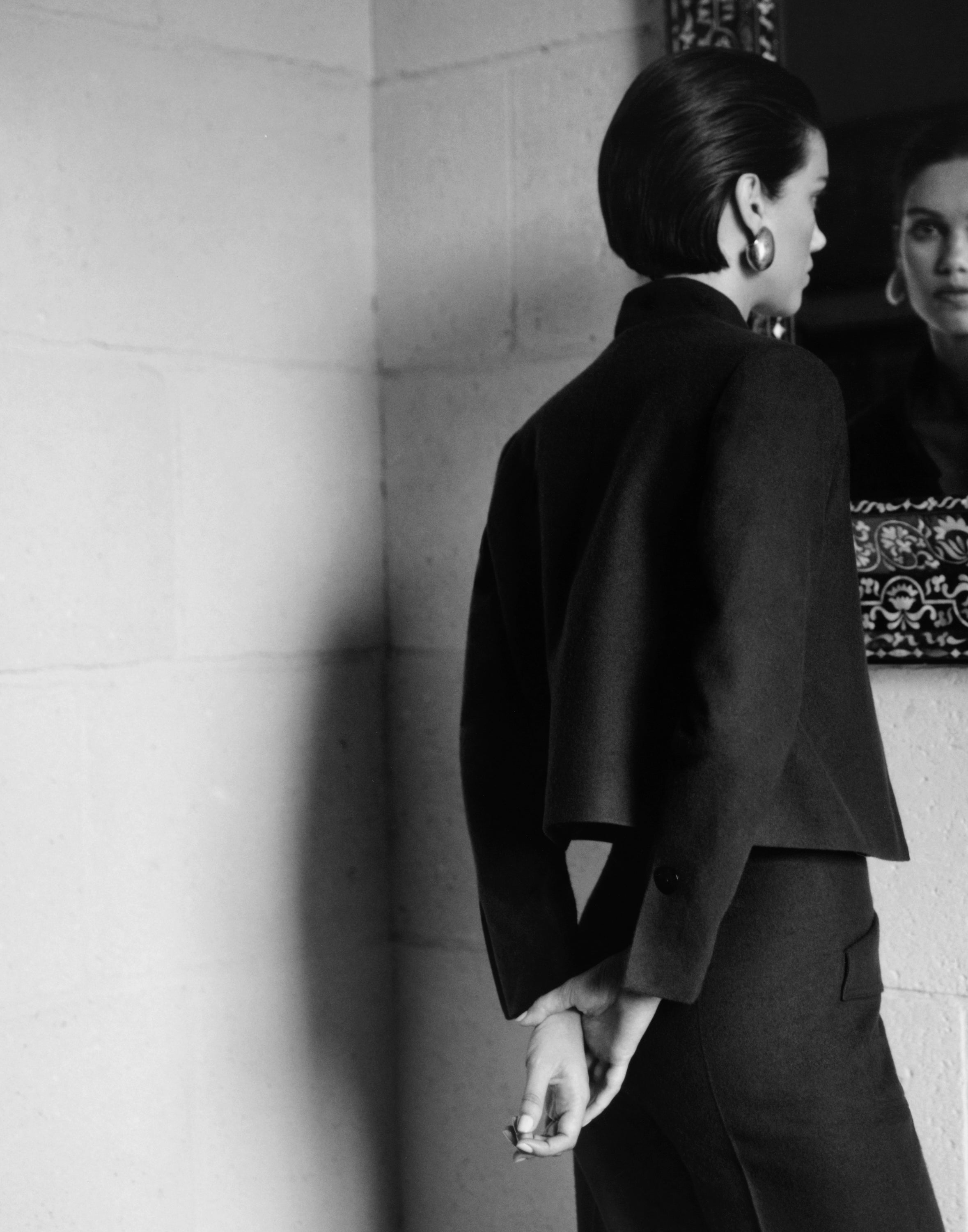 Black and white fashion photograph of a model from behind wearing a dark structured blazer and matching trousers, with a mirror reflecting her profile in a room with stone walls.