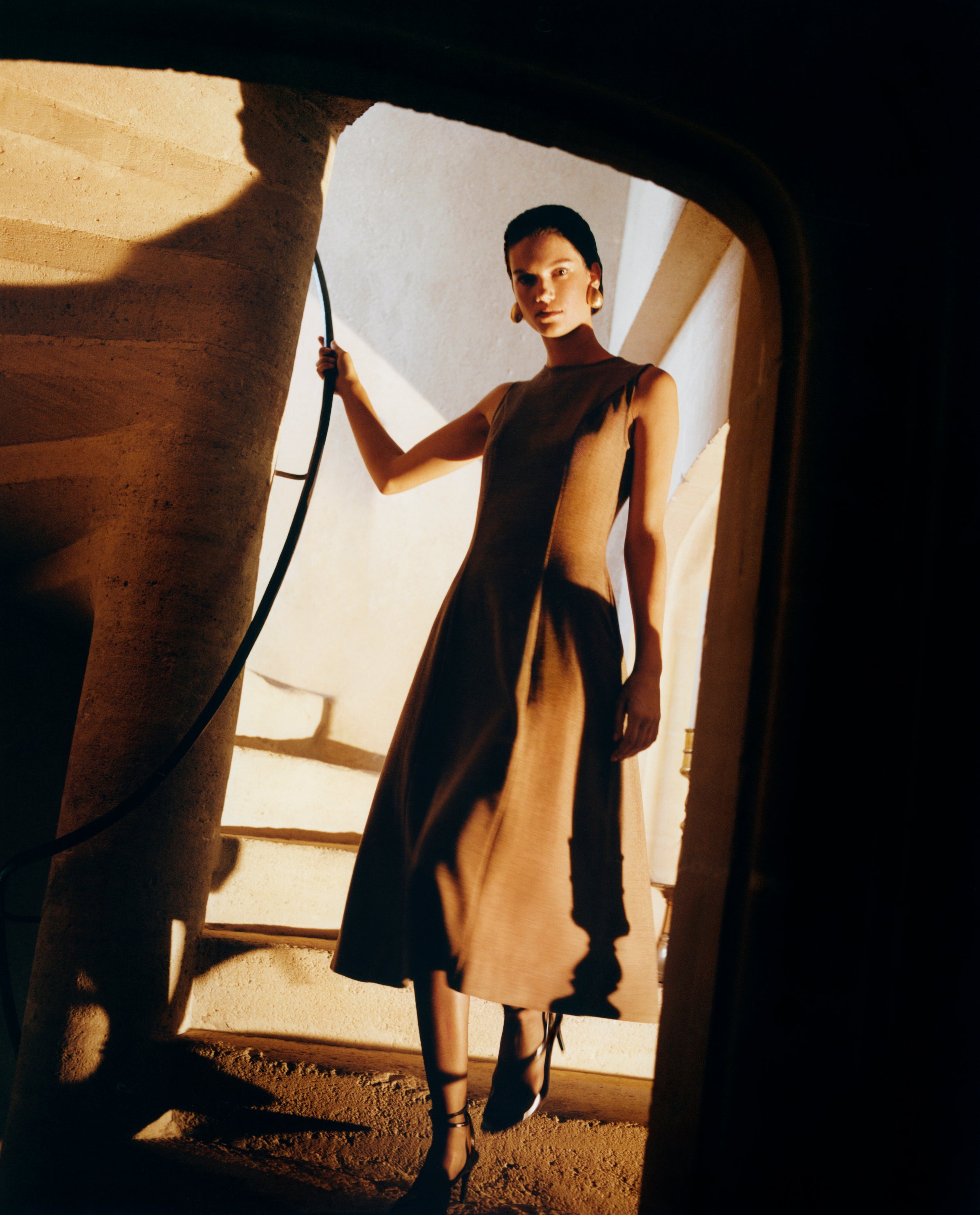 A model wearing a sleeveless, mid-calf brown dress stands in a sunlit stone interior framed by a dark archway, holding a curved black railing.