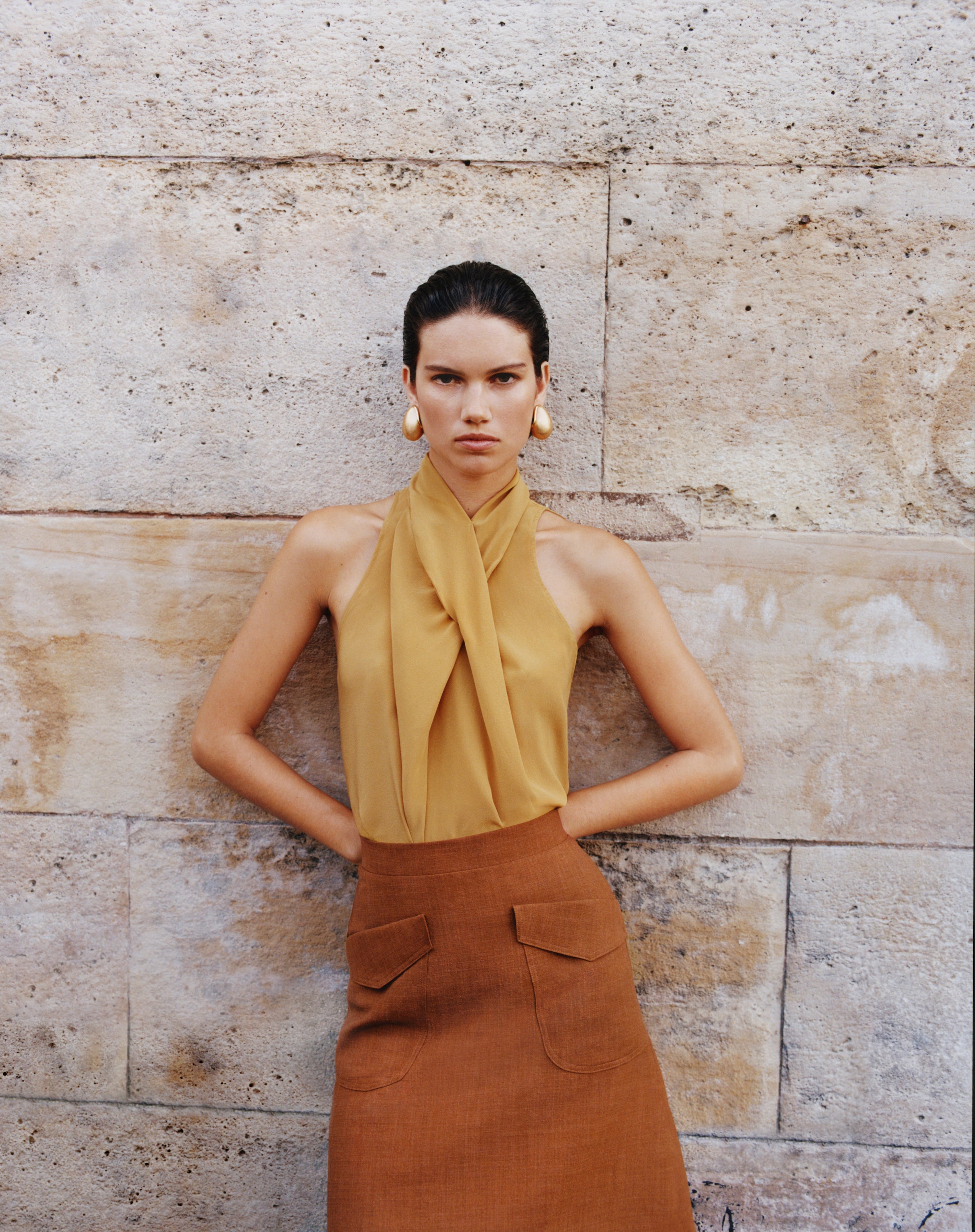 A model stands against a textured stone wall wearing a mustard yellow halter-neck top with a draped front detail and a rust linen skirt with front patch pockets.