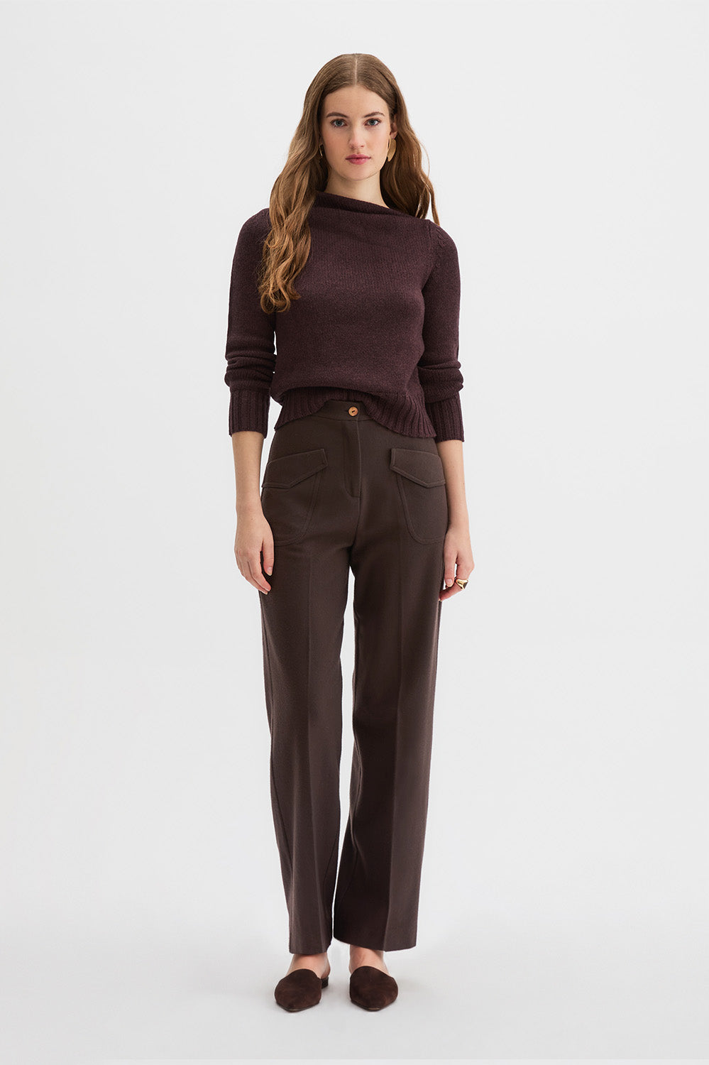 Hutton Trousers and Claude Knit. 
A woman stands against a light gray studio background wearing a dark draped sweater, dark brown wide-leg trousers, and dark brown flat shoes.