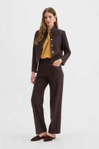 Corbu Jacket, Hutton Trousers, Ponti Top. A woman models a dark brown two-piece suit consisting of a cropped jacket with a stand collar and button details, and matching wide-leg trousers, styled with a mustard yellow blouse and dark brown pointed flats against a light gray background.