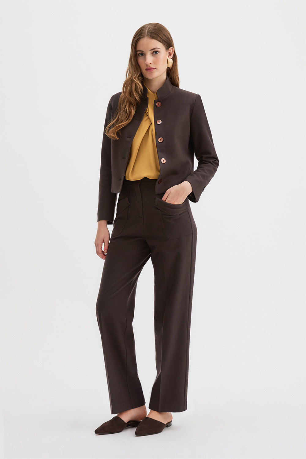 Corbu Jacket, Hutton Trousers, Ponti Top. A woman models a dark brown two-piece suit consisting of a cropped jacket with a stand collar and button details, and matching wide-leg trousers, styled with a mustard yellow blouse and dark brown pointed flats against a light gray background.