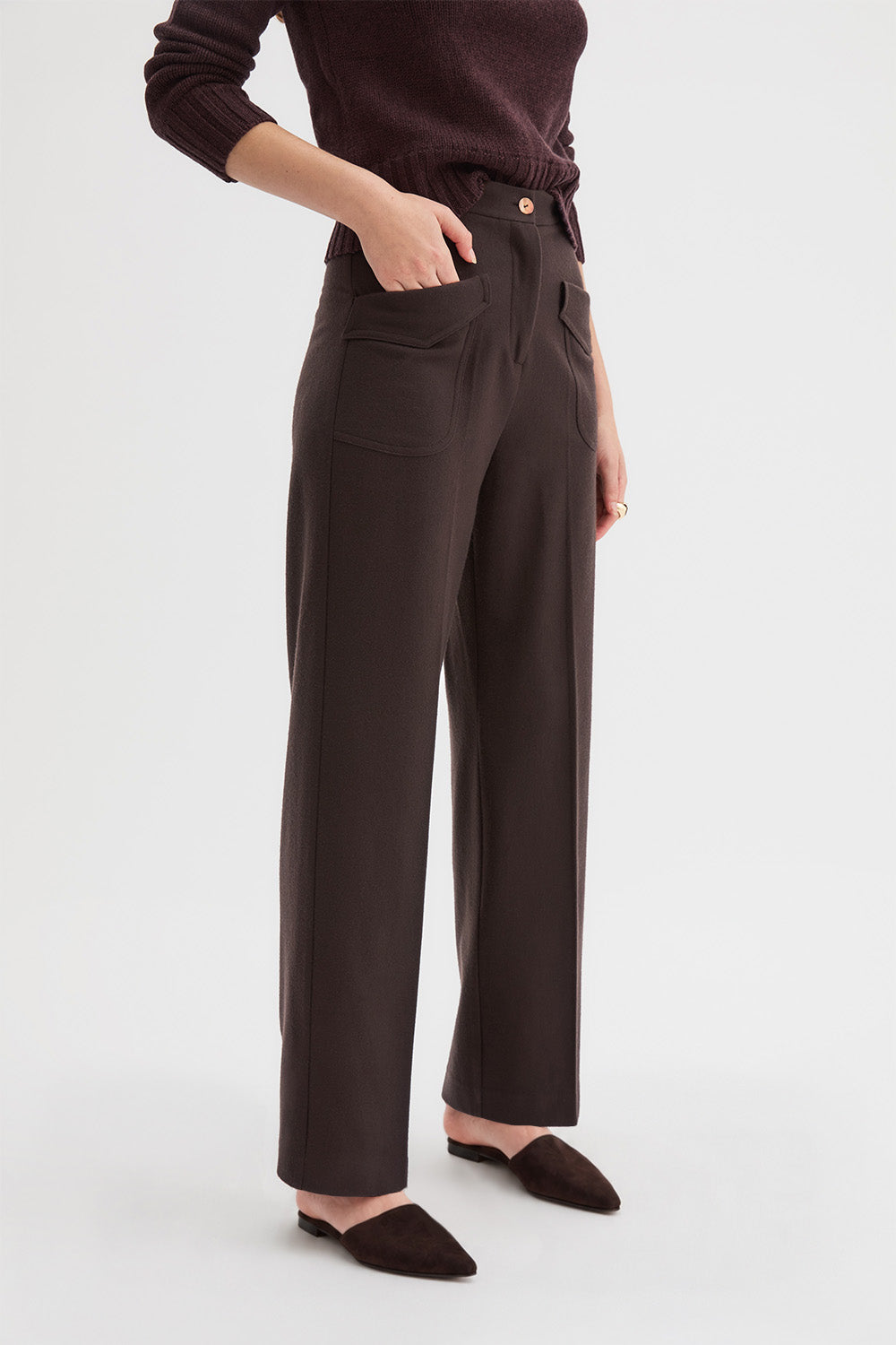 Hutton Trousers and Claude Knit. A model wearing dark brown wide-leg trousers with front patch pockets, paired with a dark brown knit top and dark brown pointed-toe mule flats, shown against a light gray studio background.