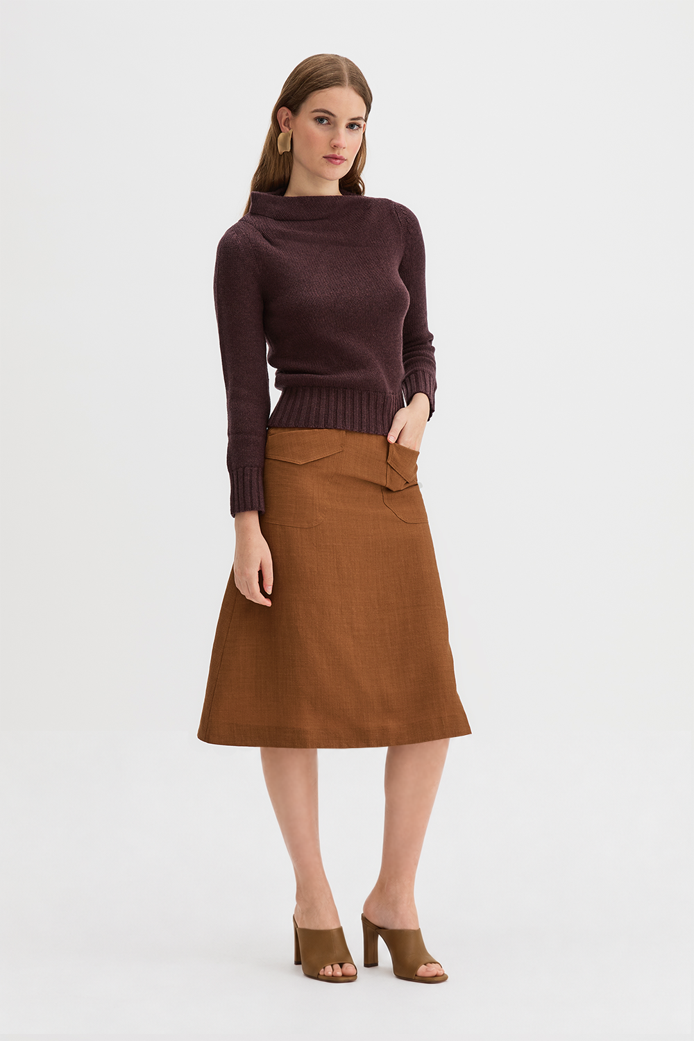 Claude Knit and Kingma A-Line Skirt. A woman modeling a dark plum ribbed knit mock-neck sweater and a brown A-line skirt, paired with brown heeled sandals, against a plain light gray studio background.
