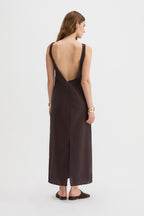 Yves Dress. A woman viewed from the back wearing a dark brown sleeveless maxi dress with a deep open back and a front slit, paired with dark brown flat shoes against a light gray studio background.