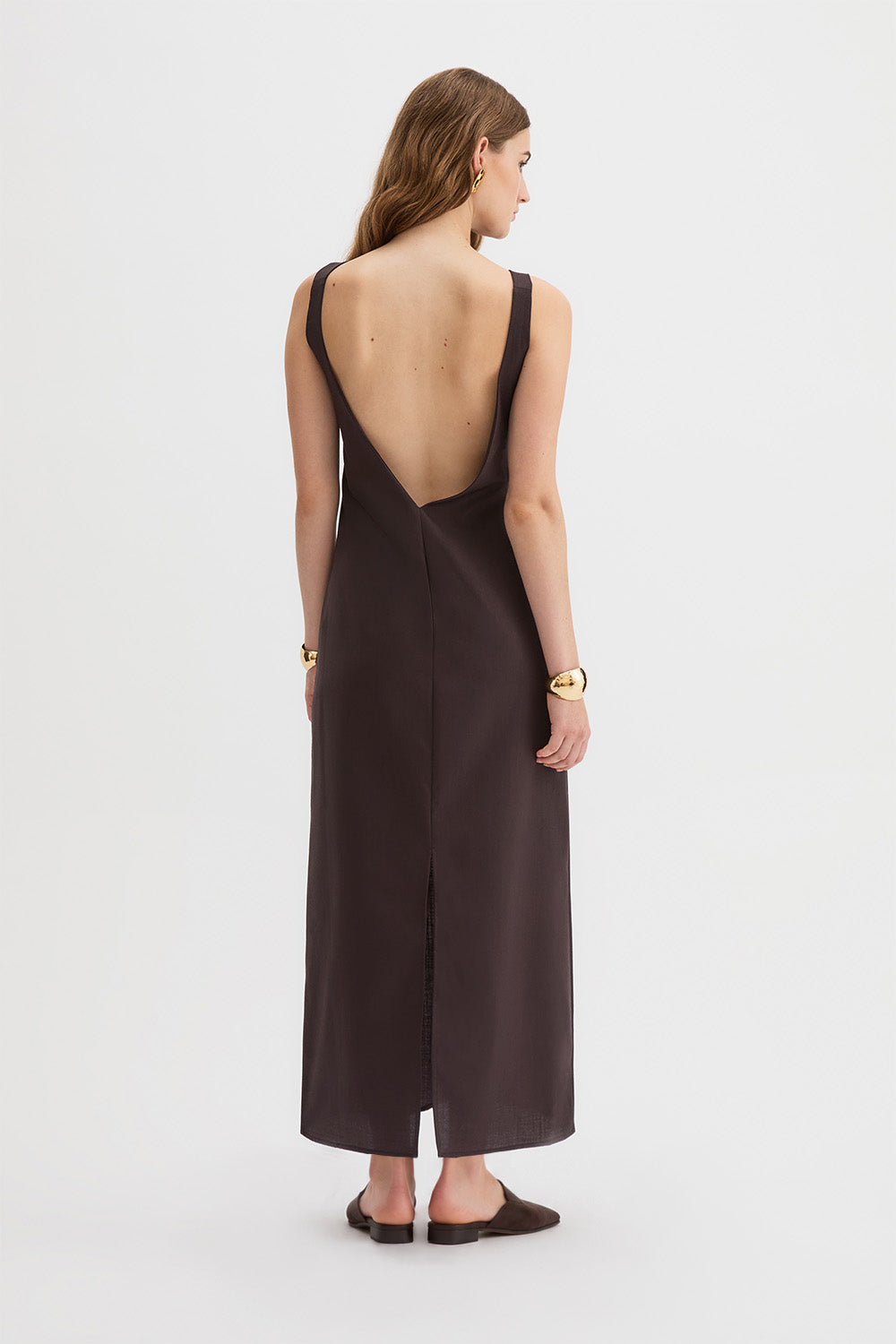 Yves Dress. A woman viewed from the back wearing a dark brown sleeveless maxi dress with a deep open back and a front slit, paired with dark brown flat shoes against a light gray studio background.