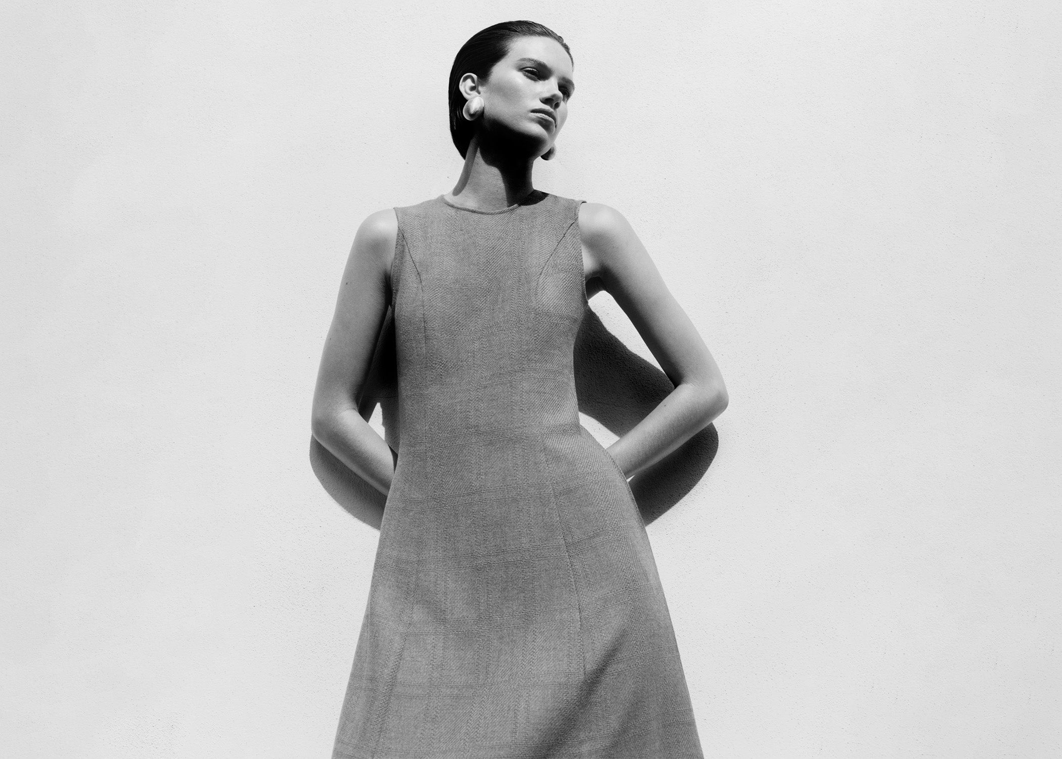 A black and white fashion photograph of a model wearing a sleeveless, high-neck, knee-length textured dress against a plain light background.