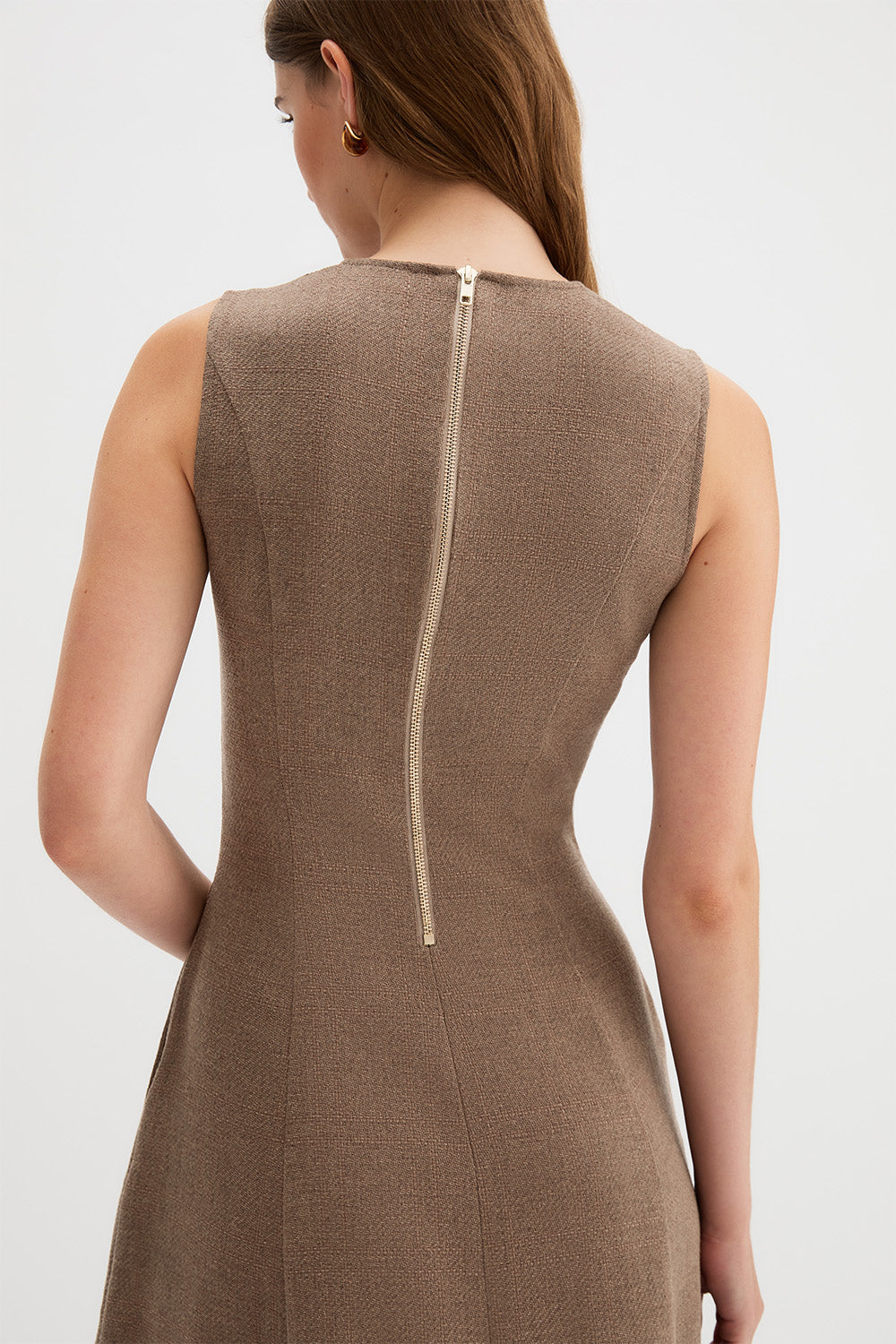 Laverne Dress, rear view of a woman wearing a sleeveless taupe-brown dress with a vertical gold-tone zipper at the back, shown against a light gray studio background.